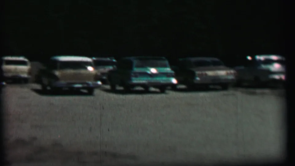 1967 home-movie frame from Farragut State Park showing a muted row of parked automobiles in a gravel lot under dark tree cover.