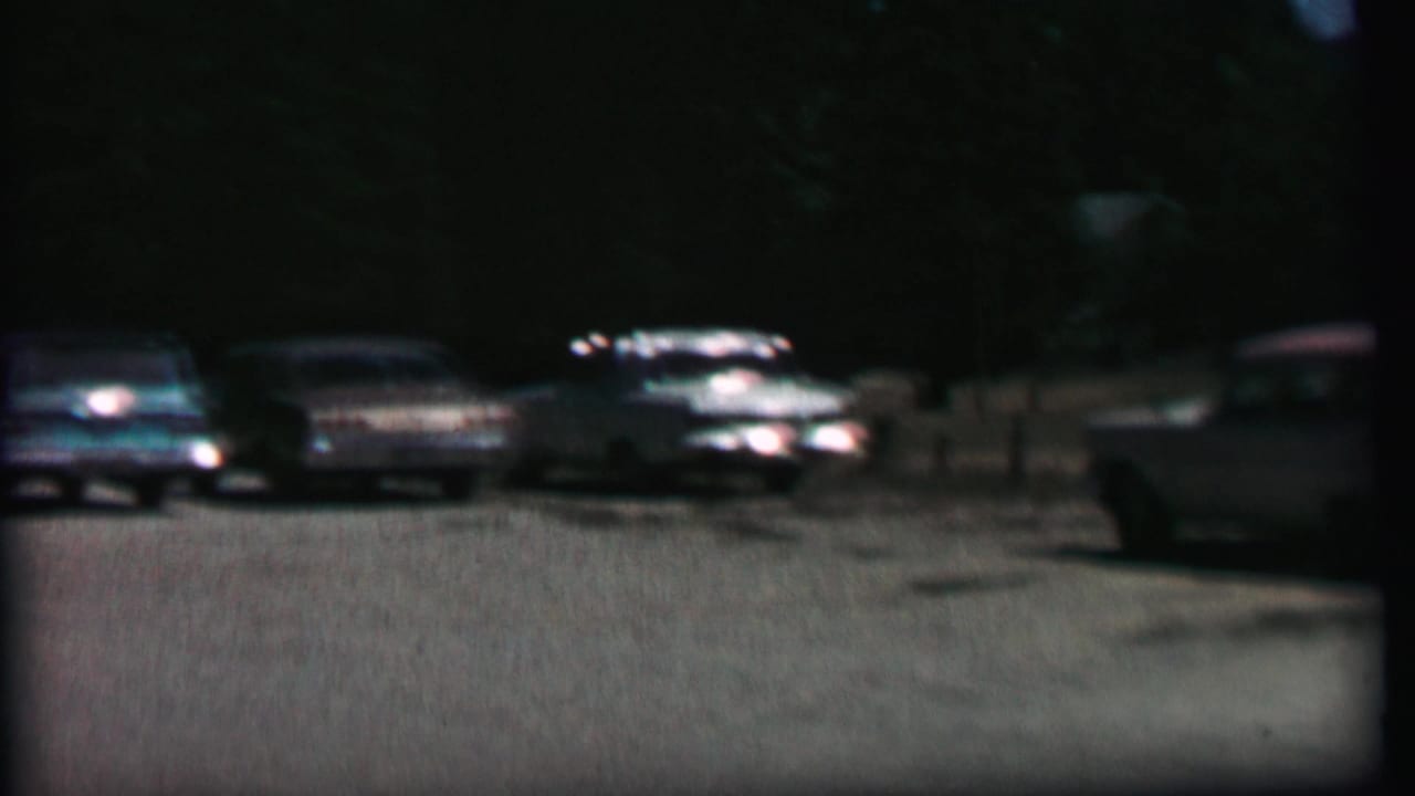 A dim 1967 home-movie frame shows several parked sedans and station wagons across a broad lot at Farragut State Park, with trees in the background.