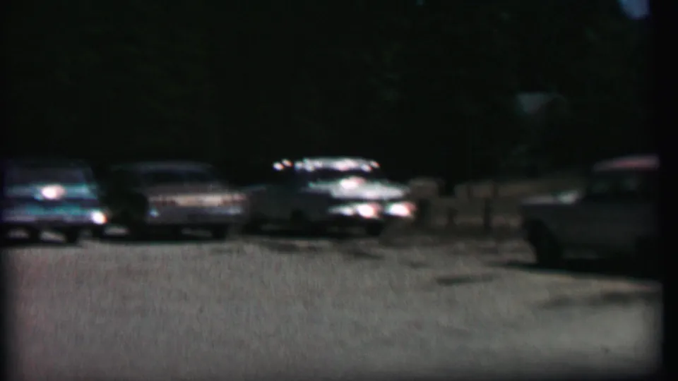 A dim 1967 home-movie frame shows several parked sedans and station wagons across a broad lot at Farragut State Park, with trees in the background.