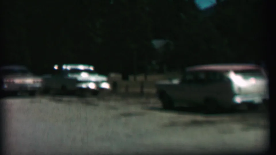 The frame shows a light-colored station wagon in the foreground of a 1967 Farragut State Park parking-lot scene, with darker cars and trees behind it.