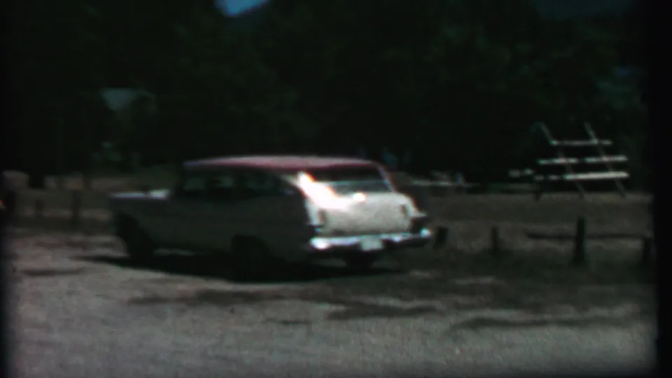 1967 Farragut State Park home-movie frame showing a white station wagon with a red roof near a split-log fence, picnic tables, gravel, and trees.