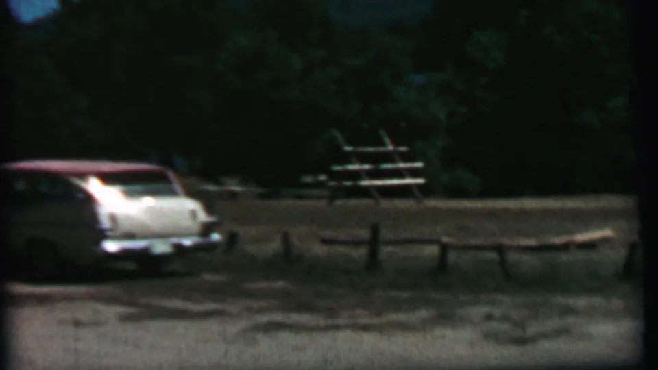Rear view of the white and red station wagon in the wooded Farragut State Park campground, with rustic fencing and picnic tables in the background.