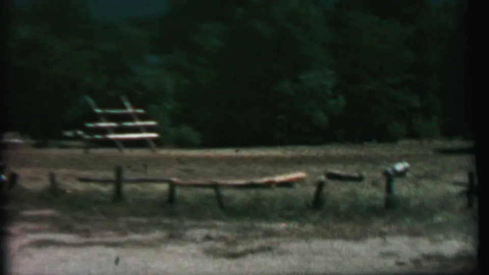 Farragut State Park campground view with split-log fencing, grass, picnic tables, gravel foreground, and dense trees after the station wagon has mostly moved out of view.