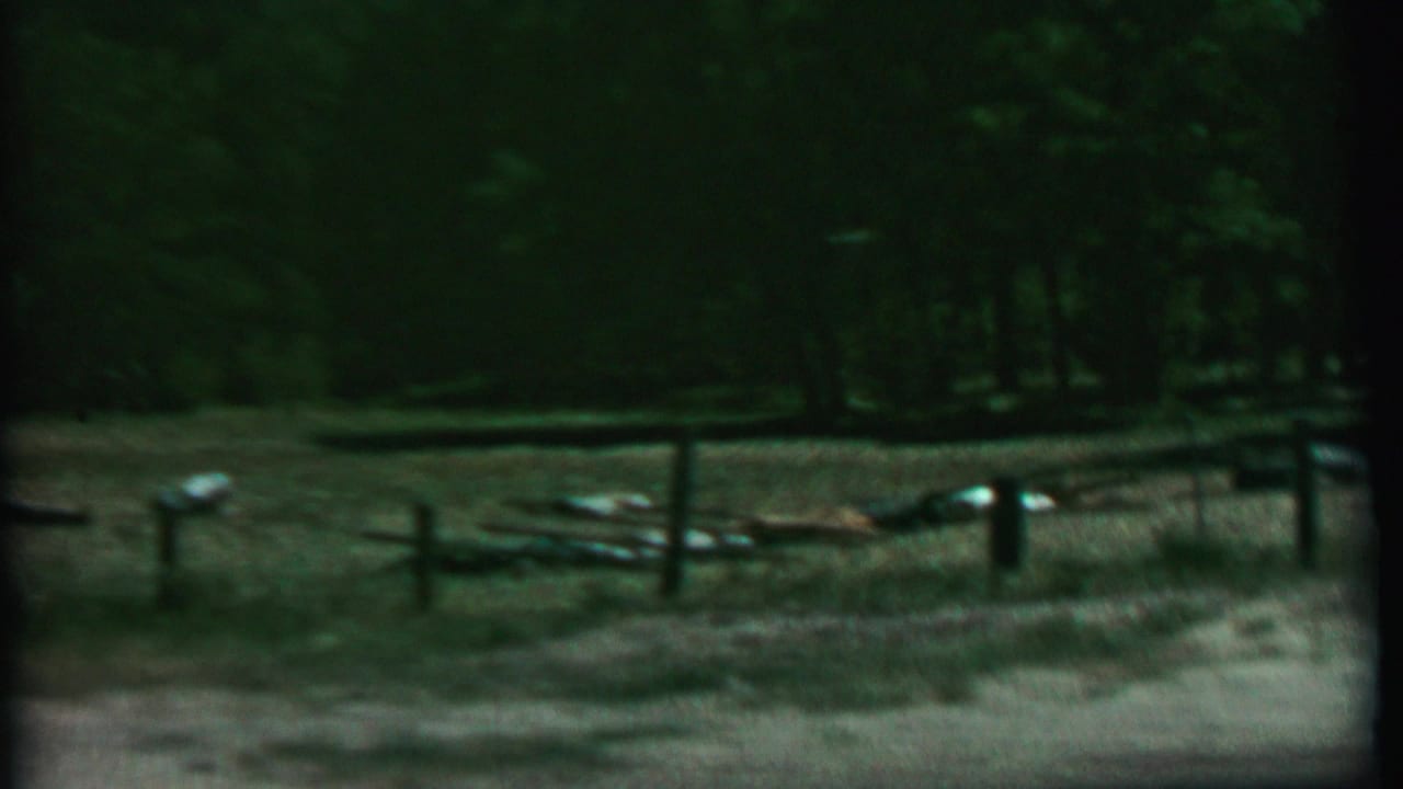 1967 8mm home-movie frame from Farragut State Park metadata showing a simple wooden fence across a dim wooded clearing with dry grass, dirt, and scattered light-colored objects.