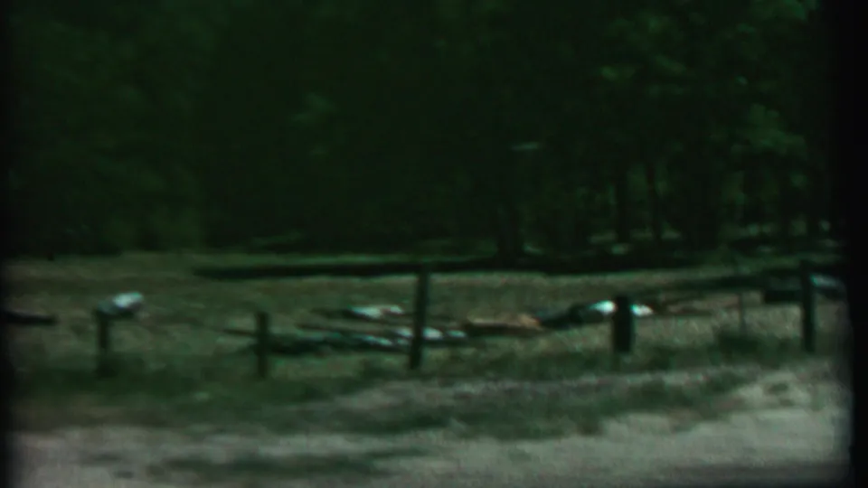1967 8mm home-movie frame from Farragut State Park metadata showing a simple wooden fence across a dim wooded clearing with dry grass, dirt, and scattered light-colored objects.
