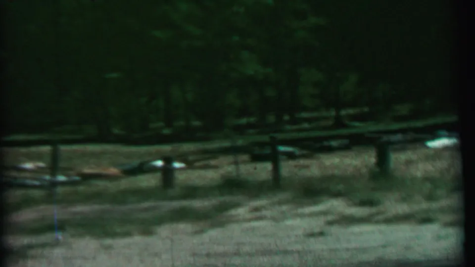 1967 Idaho park home-movie frame showing a rustic rail fence before a tree line, with small pale objects or debris scattered across the dry clearing.