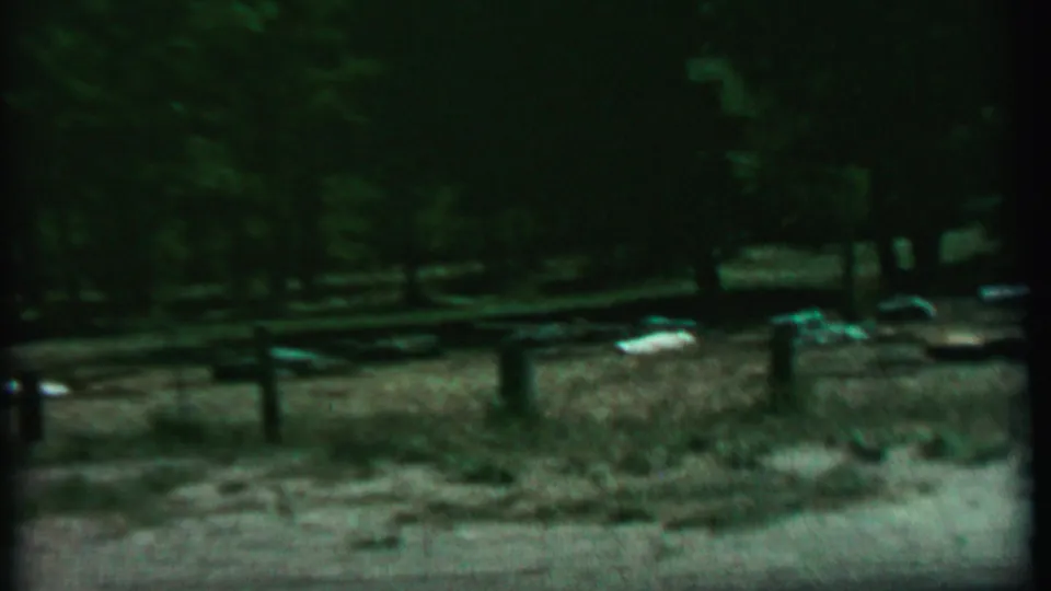Grainy 1967 8mm frame from the Farragut State Park clip showing a dim fenced enclosure, tree shadows, dry grass, dirt, and scattered light and greenish objects near the fence.