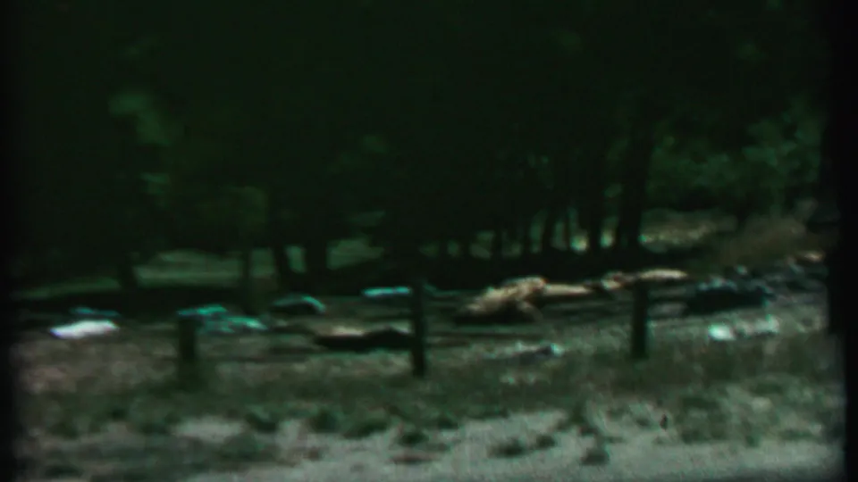 1967 home-movie frame of a wooded Farragut State Park campsite with a rustic fence, dry grass, and gear scattered near trees.