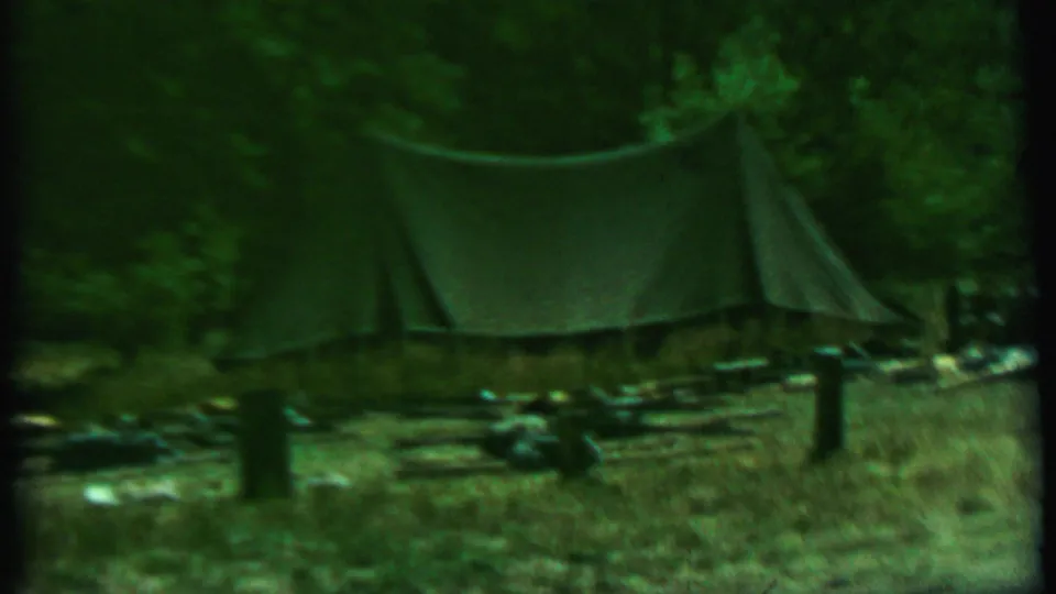 1967 Farragut State Park home-movie frame showing the canvas tent centered in a wooded campsite, with low fence posts and scattered gear in front.