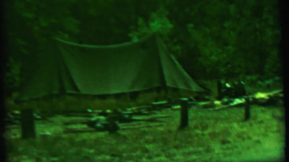 1967 8mm home-movie frame of a canvas tarp shelter at a wooded campsite in Farragut State Park, Idaho, with logs, posts, grass, and camping gear visible.