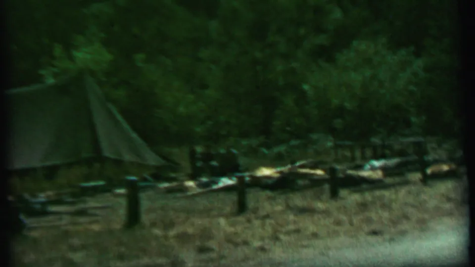 1967 8mm campsite view in Idaho with the left edge of a canvas shelter, shaded trees, rough log pieces, posts, and camping items spread along the ground.