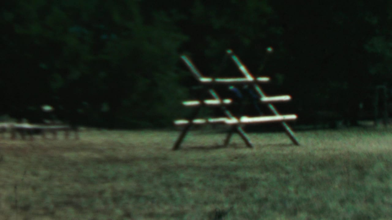 Soft 8mm frame from the supplied 1967 Farragut State Park clip showing a rustic wooden climbing structure in grass near a dark tree line.