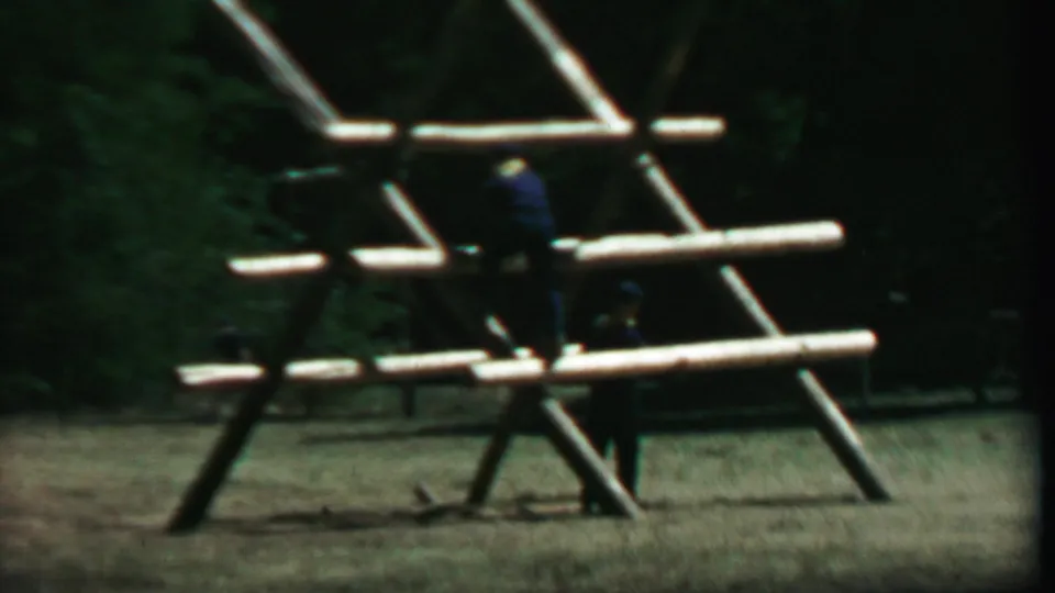 Closer 8mm view of people on and beside the large wooden climbing frame, with the grassy field and shaded trees still visible behind them.