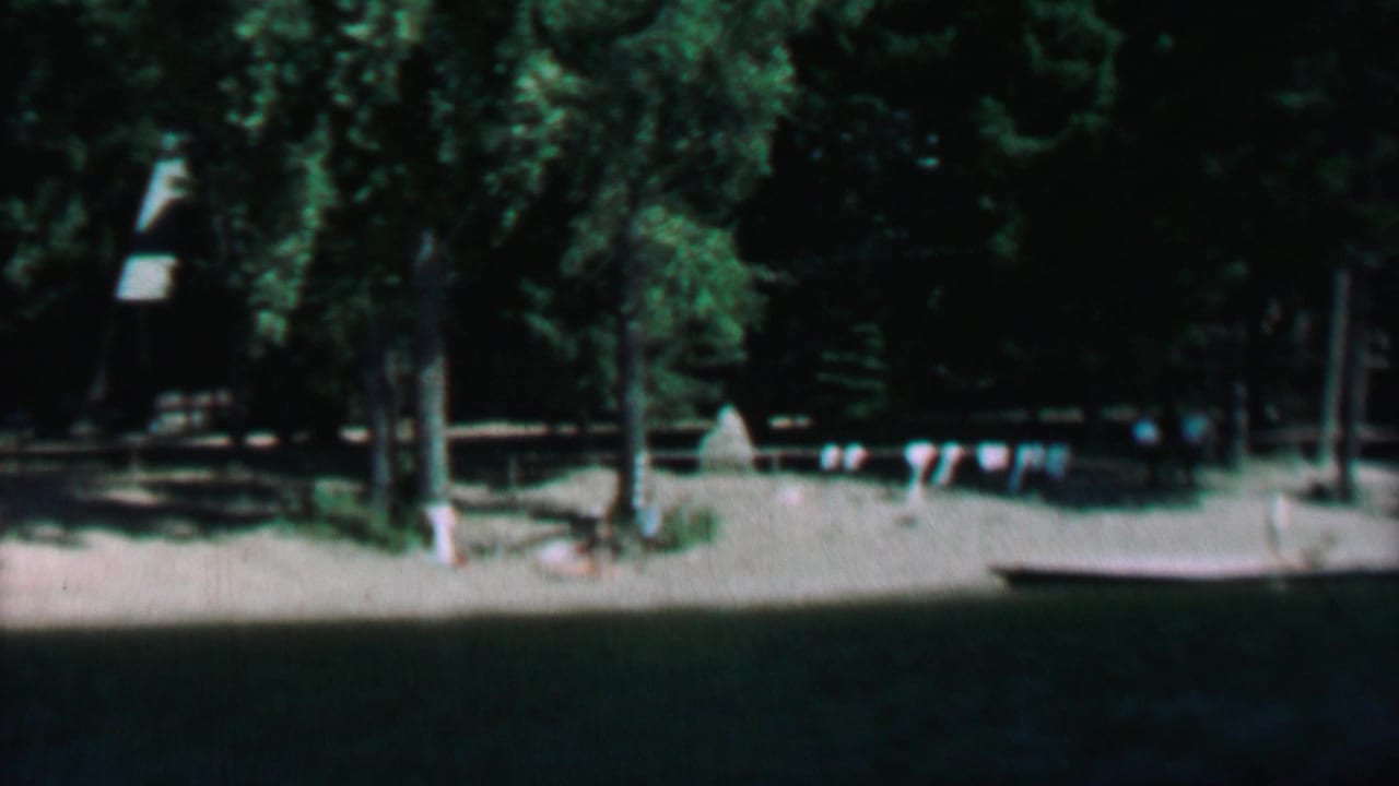 1967 8mm view at Farragut State Park, Idaho, looking across dark lake water toward a sandy shoreline with evergreen trees and small flag bunting.