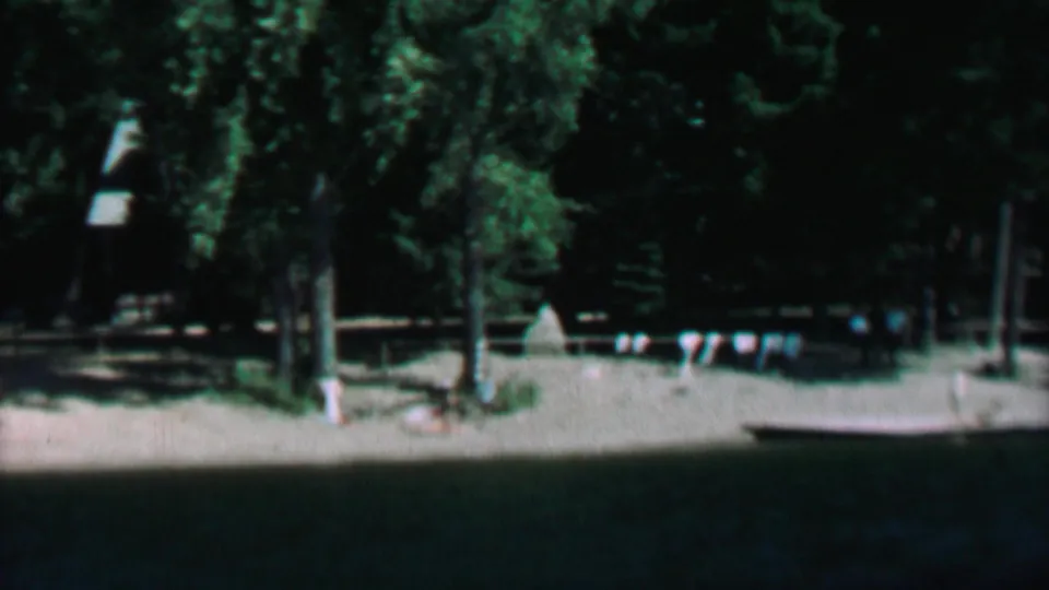 1967 8mm view at Farragut State Park, Idaho, looking across dark lake water toward a sandy shoreline with evergreen trees and small flag bunting.