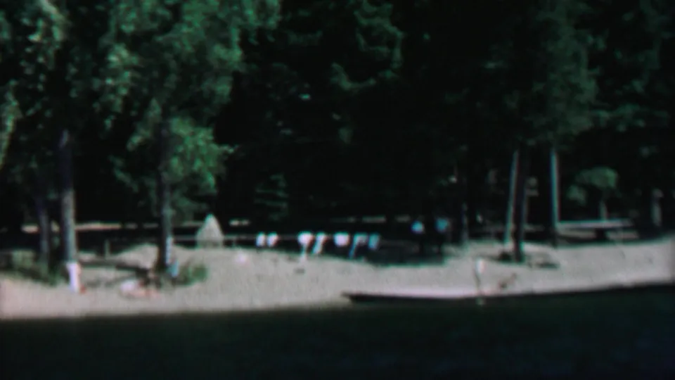 A soft 8mm frame of the Farragut State Park lakeside area, with evergreen trees, a sandy beach, flag bunting, and a low dock or platform near the water.