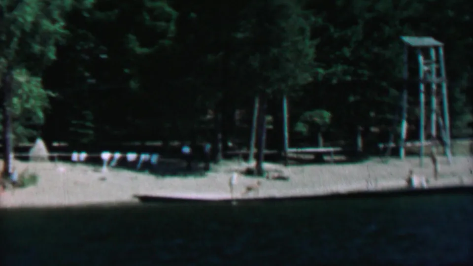 1967 lakeside home-movie frame showing a sandy shore at Farragut State Park with a dock, flag bunting, evergreen trees, and a tall wooden tower near the beach.