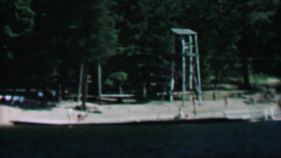 A 1967 8mm frame of Farragut State Park showing a tall wooden tower near a tree-lined lakeshore, with dock structures, flags, and dark water in the foreground.