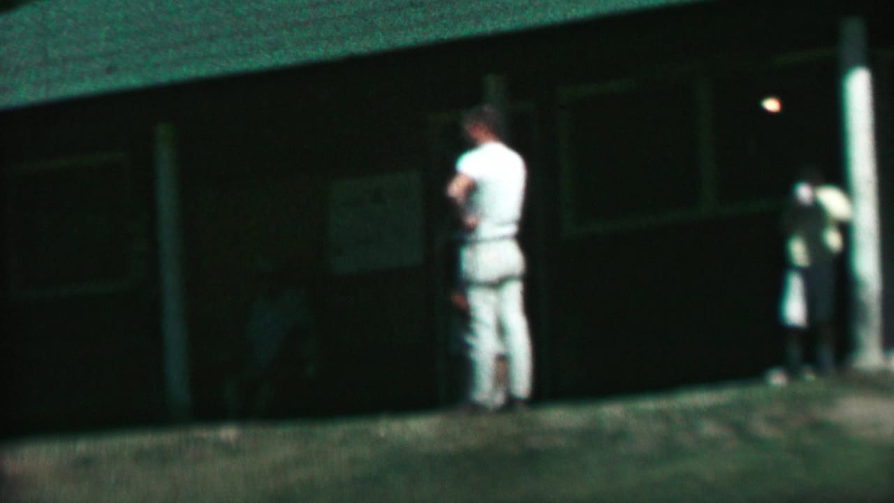 1967 home-movie frame at Farragut State Park showing a man in white clothing near a dark wooden park building with grass and trees around it.