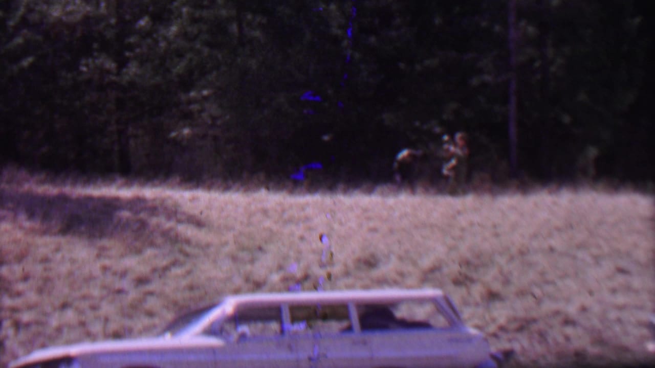 Supported by the clip metadata, this 1967 Farragut State Park frame shows a light-colored vintage station wagon in the foreground and small distant figures near the wooded edge of a grassy field.