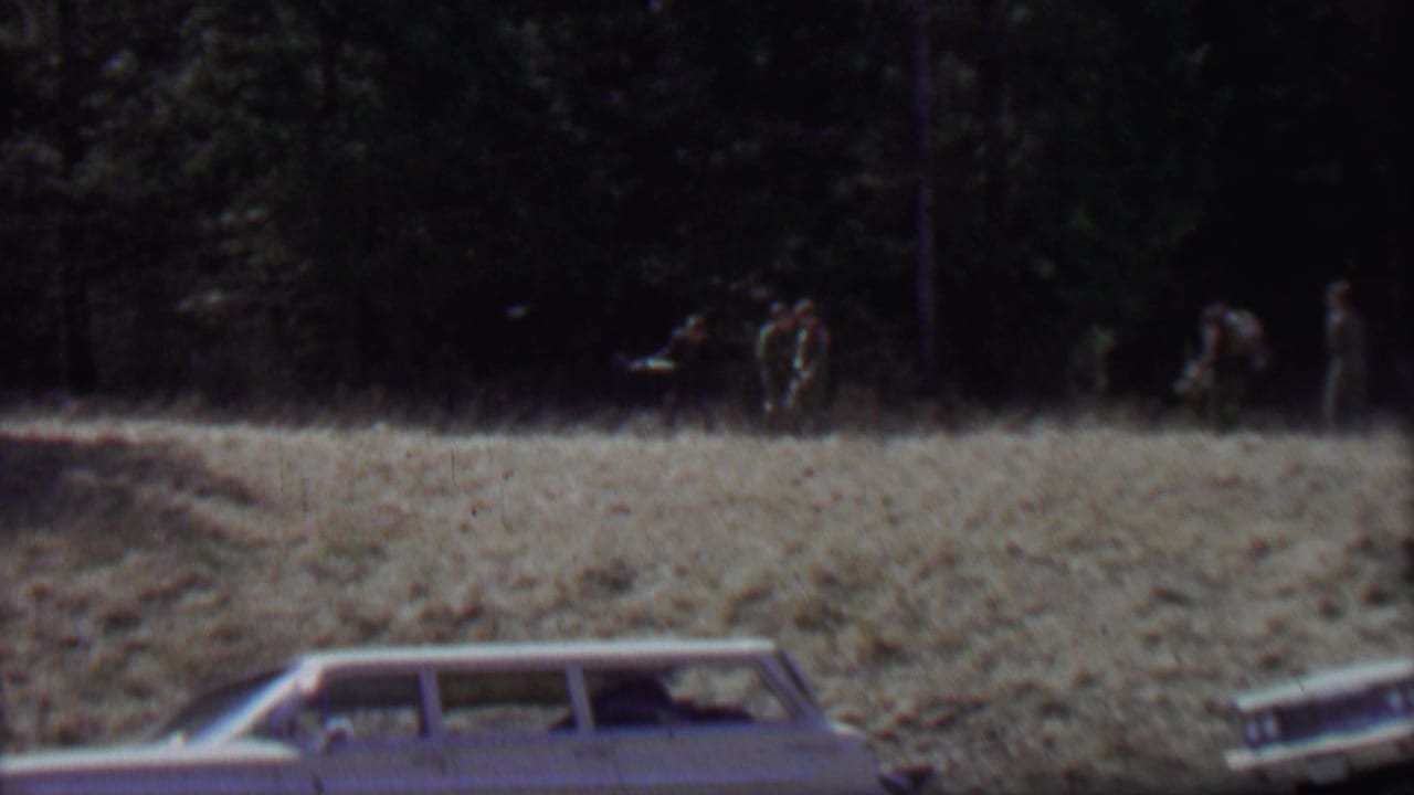 Station wagon in foreground with several distant people walking along the edge of a wooded field.