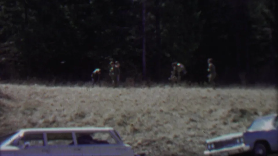 This 1967 Idaho park home-movie frame shows small figures gathered or walking near the dark tree line, with the roof and windows of a vintage station wagon visible in the foreground.