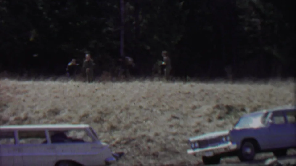 Supported by the clip metadata, this Farragut State Park frame shows two vintage cars at the field edge and a few distant people standing or moving near the wooded background.