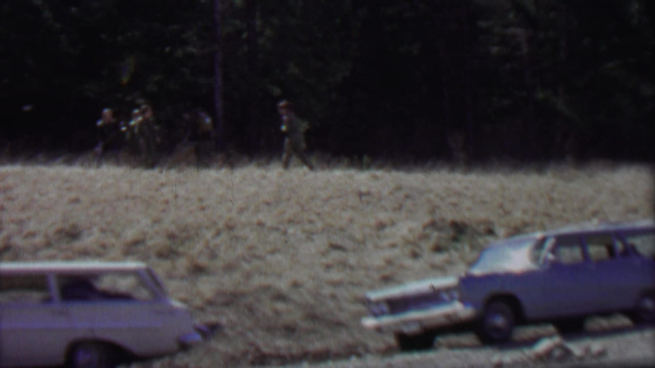 1967 8mm frame from Farragut State Park, Idaho, showing classic station wagons along a roadside and distant men walking near evergreen trees.
