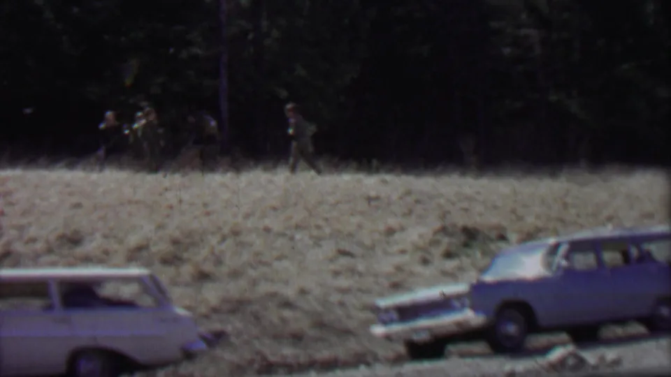 1967 8mm frame from Farragut State Park, Idaho, showing classic station wagons along a roadside and distant men walking near evergreen trees.