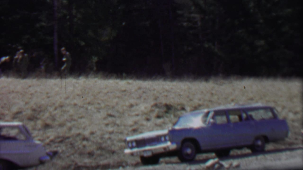 A blue station wagon and a white station wagon sit near the road while distant men walk across a dry field at Farragut State Park in the 1967 footage.