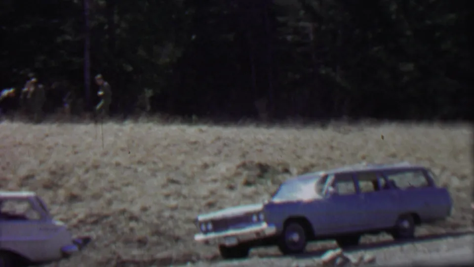 A blue station wagon and a white station wagon sit near the road while distant men walk across a dry field at Farragut State Park in the 1967 footage.