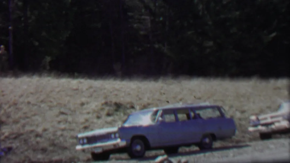 The frame centers on a blue 1960s station wagon at the roadside, with another wagon partly visible and a dry field leading back to evergreen trees.