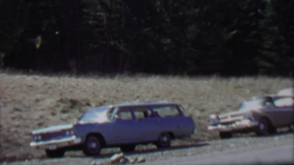 A blue station wagon remains parked along the road with a partial white vehicle nearby, set against the dry field and evergreen backdrop identified in the clip metadata as Farragut State Park.