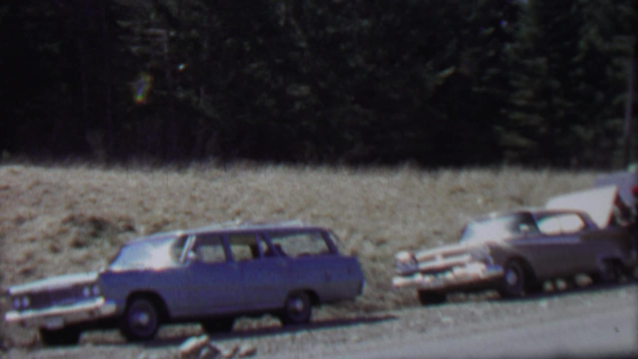 1967 home-movie frame from the supplied Farragut State Park clip showing a blue station wagon and light-colored sedan parked along a gravel roadside below a forested slope.