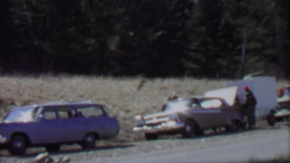 1967 Farragut State Park home-movie frame showing parked 1960s cars, a white trailer or raised panel, and adults standing near the vehicles on a gravel roadside.