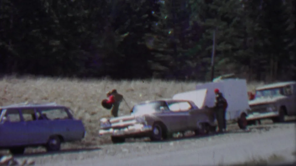 1967 home-movie frame from Farragut State Park showing an adult in red walking near parked cars, with another person beside a white trailer or panel along the gravel road.