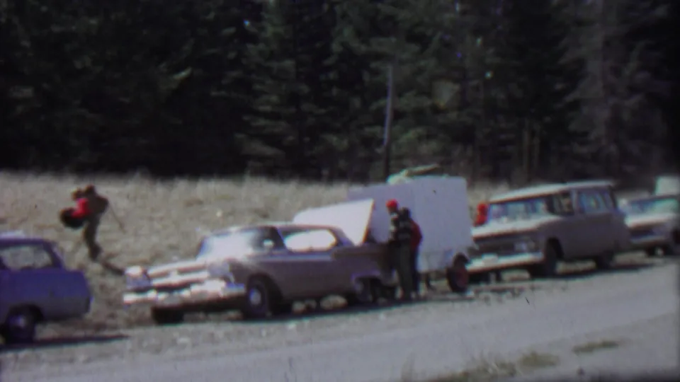1967 Farragut State Park frame showing adults near a white trailer or raised panel, with a sedan and station wagons parked along a gravel roadside below dense trees.