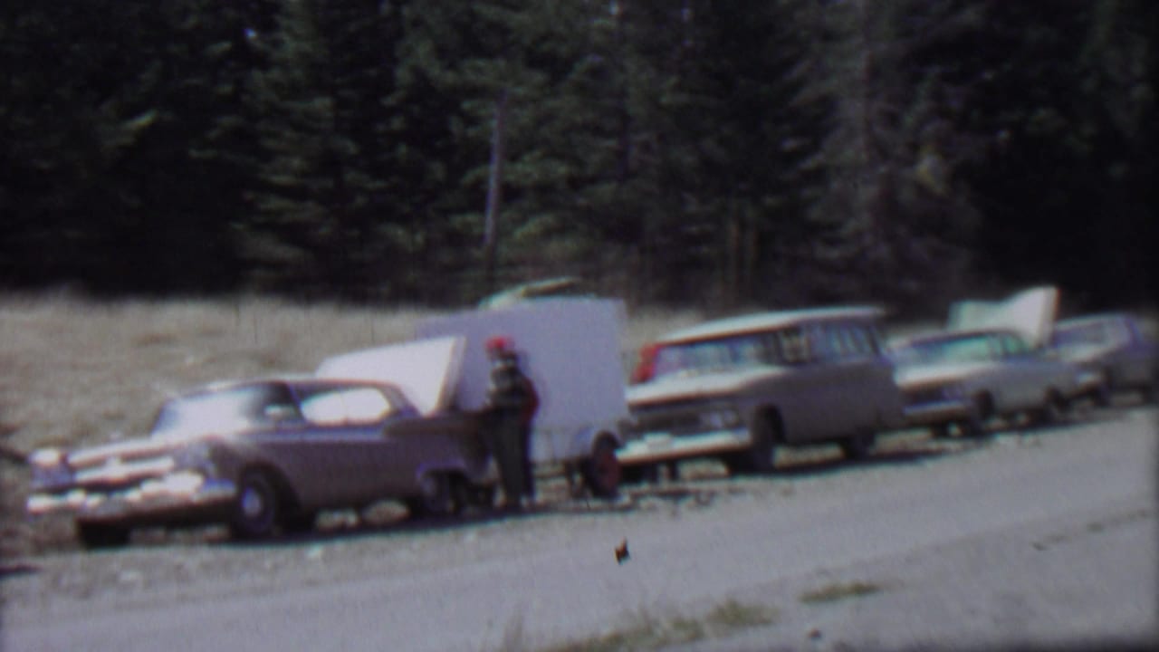 1967 Farragut State Park home-movie frame showing several older cars along a roadside, a white camper trailer or panel, and a man in a red cap near dense evergreens.