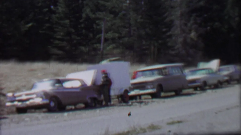 1967 Farragut State Park home-movie frame showing several older cars along a roadside, a white camper trailer or panel, and a man in a red cap near dense evergreens.