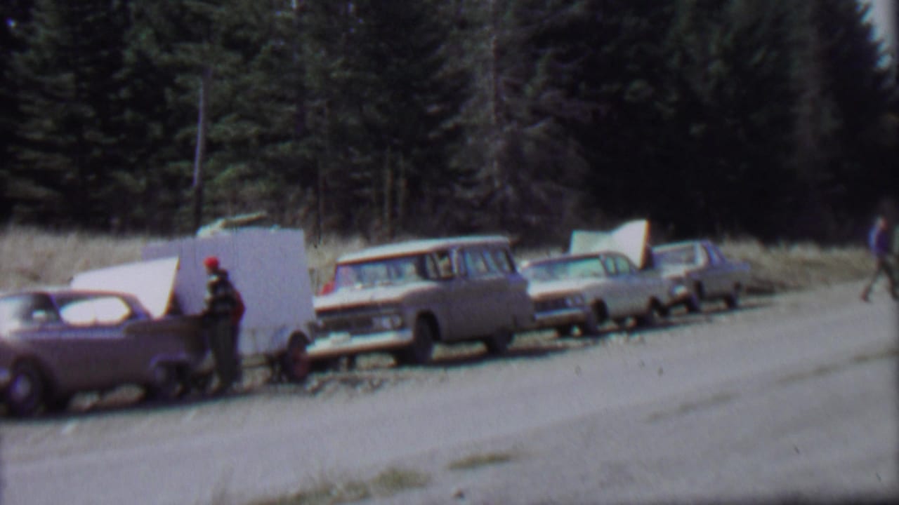 1967 Idaho park roadside view with a line of 1960s-era cars, a white camper trailer or panel, and a person standing near the vehicles under a bright sky.