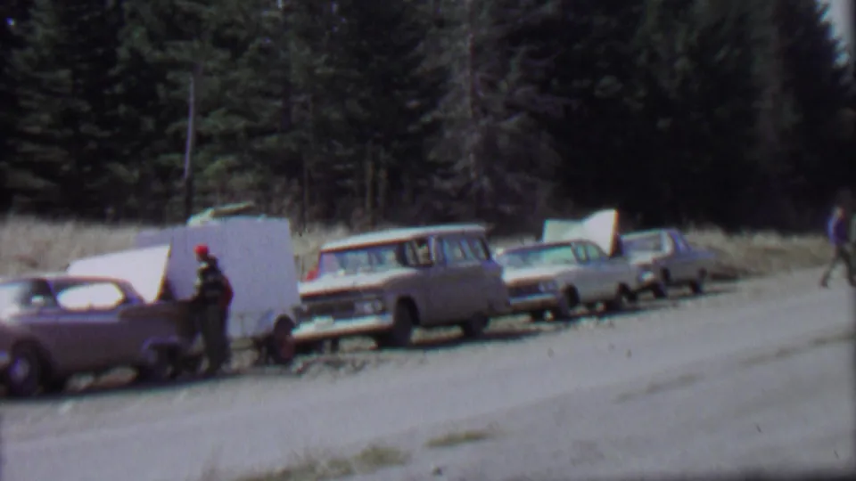 1967 Idaho park roadside view with a line of 1960s-era cars, a white camper trailer or panel, and a person standing near the vehicles under a bright sky.