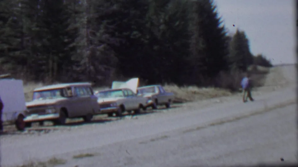 Home-movie frame from the 1967 Farragut State Park clip showing parked cars with open hoods along a road and a distant walker near the wooded shoulder.