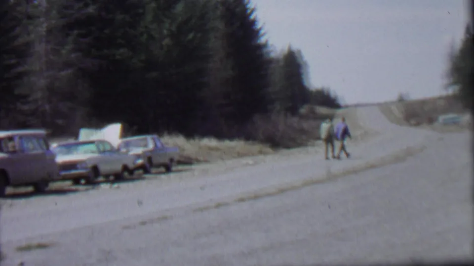1967 Farragut State Park roadside frame with two distant people walking, several parked cars along the shoulder, and dark evergreen trees framing the road.