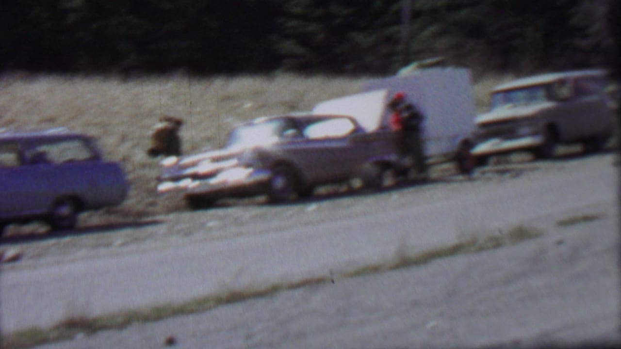 1967 Farragut State Park home-movie frame showing vintage cars near a gravel roadside, with people gathered beside a sedan whose trunk appears open.