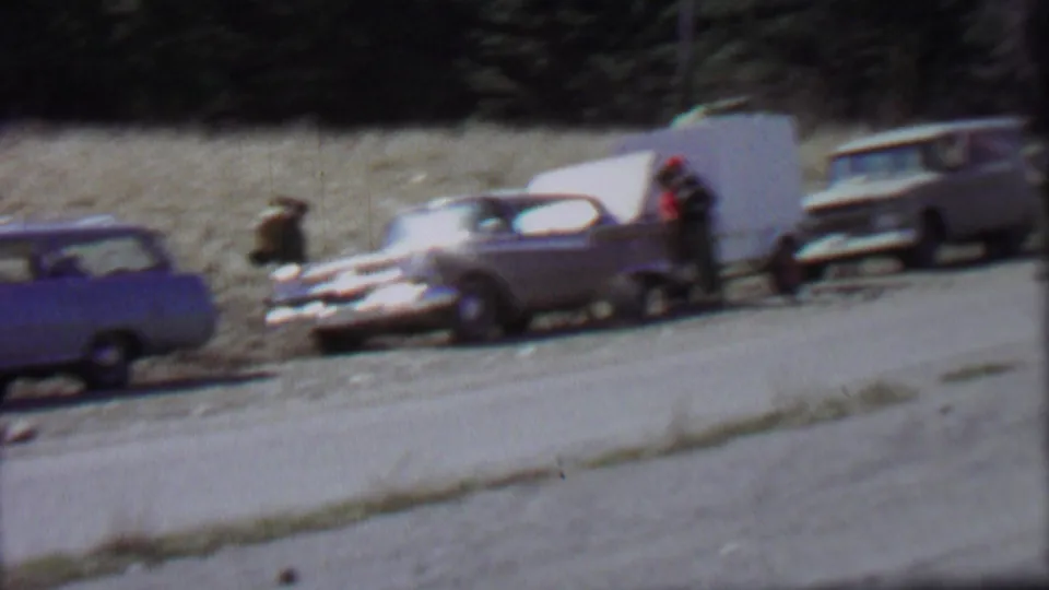 1967 Farragut State Park home-movie frame showing vintage cars near a gravel roadside, with people gathered beside a sedan whose trunk appears open.