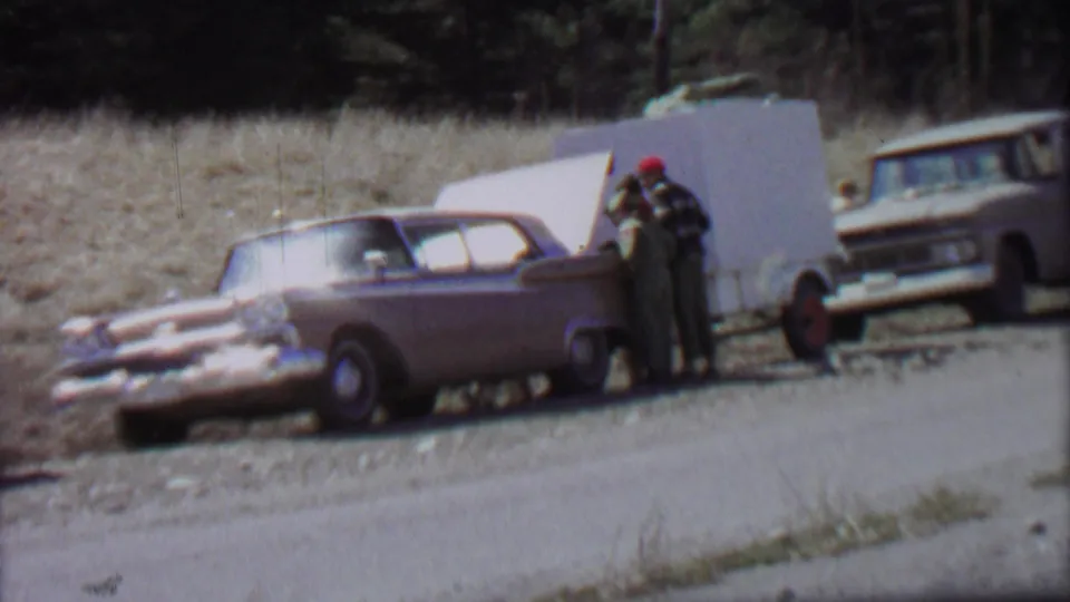 1967 Farragut State Park home-movie frame with two people beside a parked sedan and open trunk, backed by dry grass, trees, a trailer, and another vintage car.