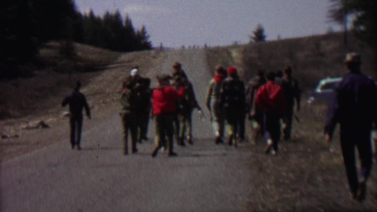 In supplied 1967 Farragut State Park 8mm footage, a group of scouts or outdoor participants walk away from the camera along a rural road with pine trees and rocks nearby.