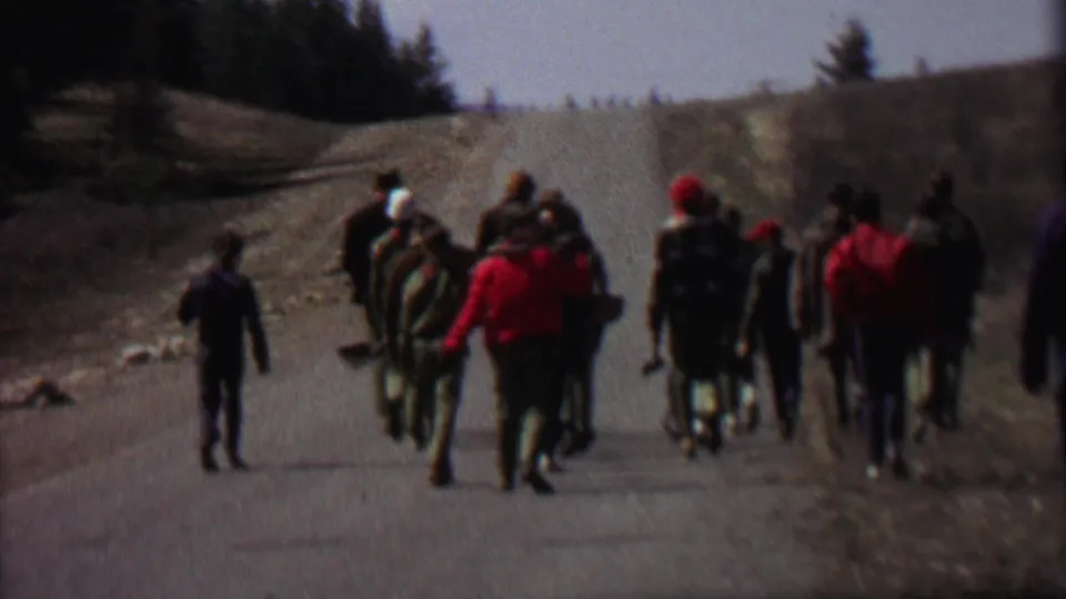 The group continues down the rural road in the supplied Idaho park footage, with several participants carrying packs and tools and a light-colored station wagon visible near the bend.