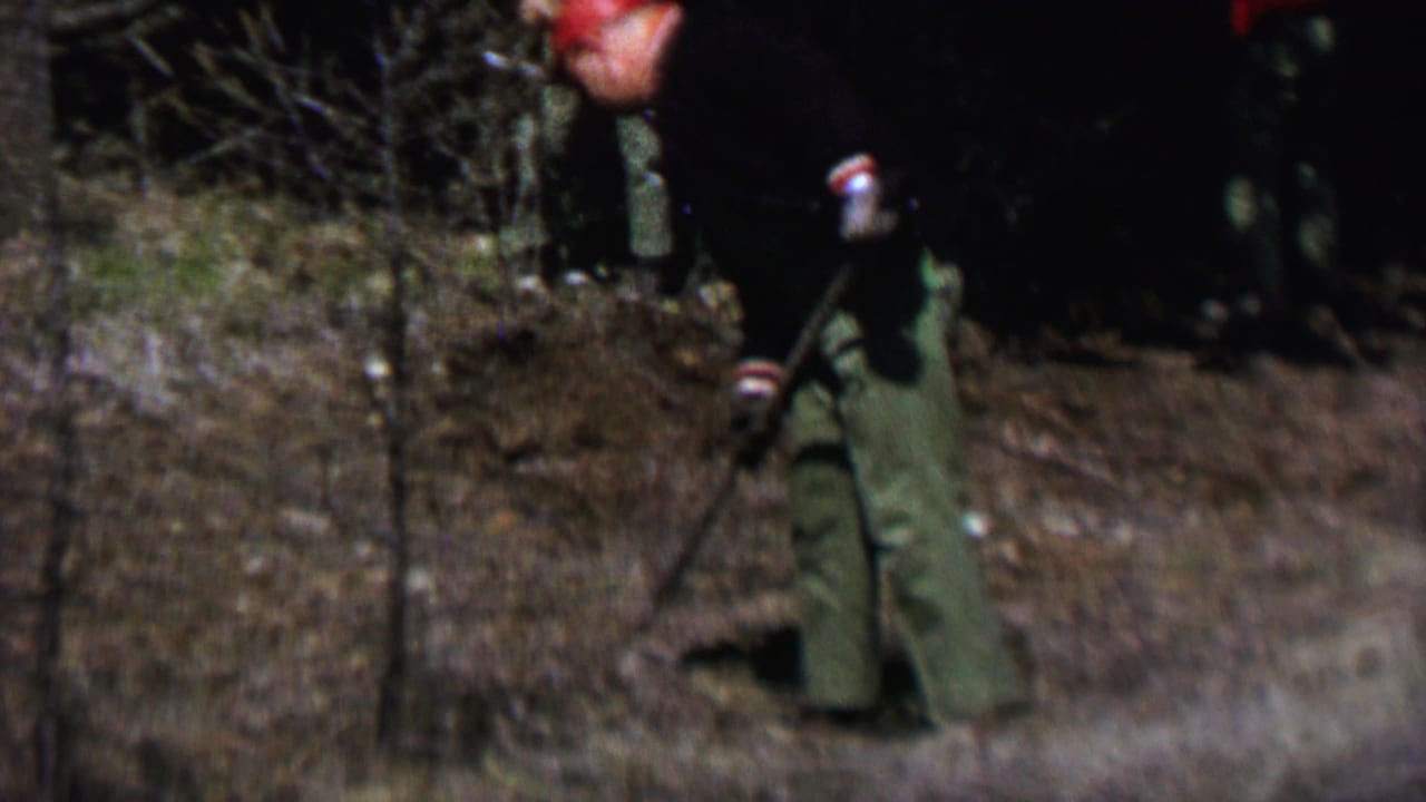 In 1967 Farragut State Park footage, a man in a red cap, dark jacket, and green trousers clears dead vegetation on a brushy woodland slope.