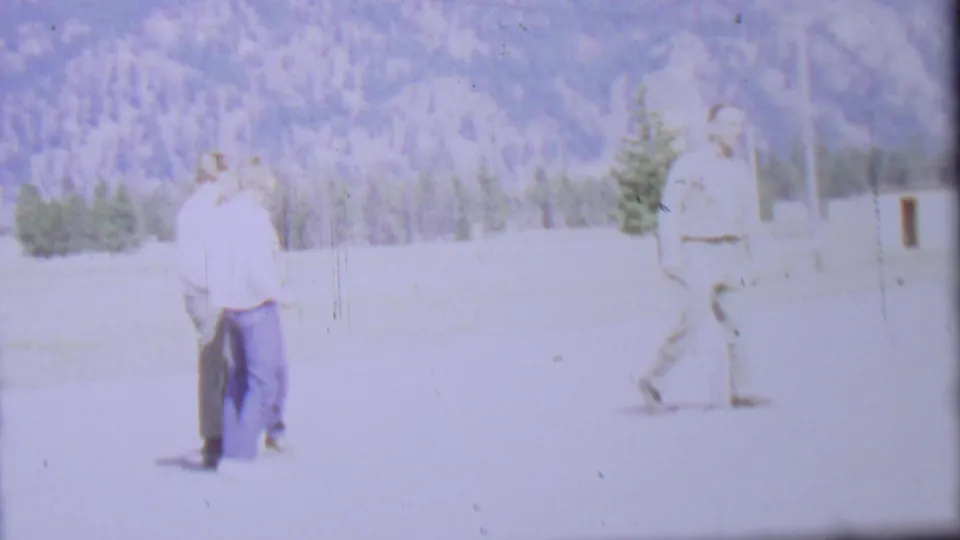 1967 8mm frame from Farragut State Park metadata showing two adult men in a sunlit field as another man walks across the right side with trees and mountains in the background.
